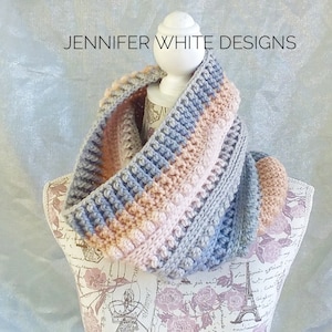 Könnte beinhalten: Ein gehäkelter Infinity-Schal mit einem strukturierten Stichmuster in Grautönen, Pfirsich und Rosa. Der Schal wird auf einer Puppe mit einem Blumenmuster präsentiert.