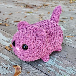 Könnte beinhalten: Ein Plüsch-Häkeltier in Form einer Katze in rosa Farbe mit schwarzen Augen. Die Katze sitzt auf einer Holzoberfläche.