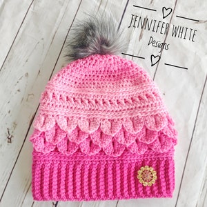 May include: A pink and white crocheted hat with a faux fur pom pom on top. The hat has a scalloped edge and a sparkly embellishment on the side. The hat is labeled "Jennifer White Designs".