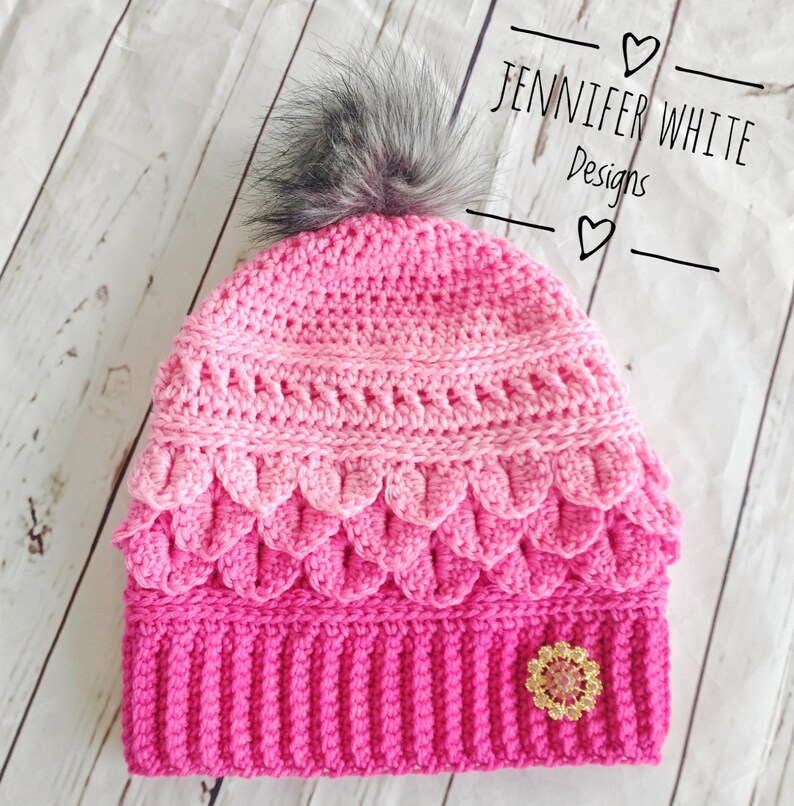 May include: A pink and white crocheted hat with a faux fur pom pom on top. The hat has a scalloped edge and a sparkly embellishment on the side. The hat is labeled "Jennifer White Designs".