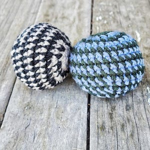 May include: Two spherical, hand-crocheted balls. One ball features a tight, alternating pattern of black and white yarn. The second ball displays a textured, interwoven pattern of light blue and dark forest green yarn, both showing a dense stitch.