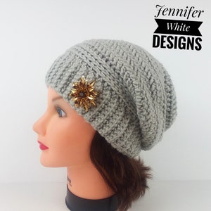 Puede incluir: Boina de punto de ganchillo gris con un broche de flor dorada. La boina es holgada y tiene un patrón de punto texturizado. Jennifer White Designs.