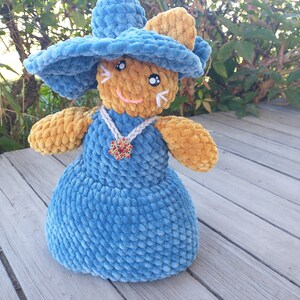 May include: A crocheted plush toy in the shape of a cat wearing a blue hat and a blue dress with a red and gold pendant. The cat has a brown face and large black eyes.