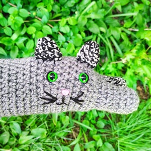May include: A handmade gray crochet cat toy with black and white ears, green eyes, a pink nose, and black whiskers. The cat is lying on a bed of green leaves and grass. The toy is soft and plush.