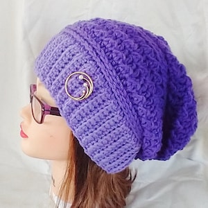 Puede incluir: Boina de punto de ganchillo morado con un adorno de oro y perlas.