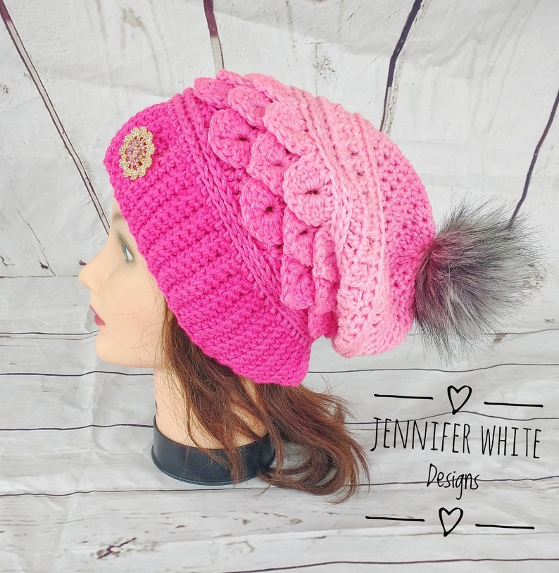 May include: A pink and white ombre crochet beanie with a faux fur pom pom and a sparkly embellishment. The beanie is on a mannequin head with long brown hair.