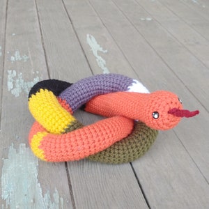 May include: A crocheted snake toy in orange, yellow, purple, and green. The snake is curled into a circle and has a red nose.