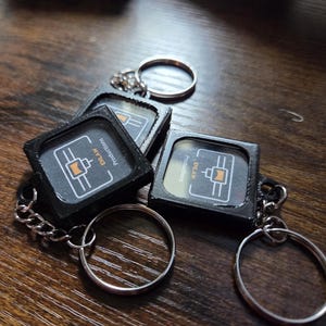Puede incluir: Tres llaveros cuadrados negros con anillas de metal plateado. Cada llavero presenta un gráfico central de un icono de boquilla de impresora 3D en naranja y blanco con el texto ENLAW Productions impreso en blanco y naranja sobre fondo oscuro.