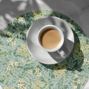May include: A green and white floral patterned round coaster with a white saucer and a cup of coffee on top.