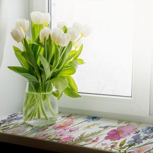 Puede incluir: Un jarrón lleno de tulipanes blancos se encuentra en un alféizar de ventana con una superficie de patrón floral. El jarrón es de vidrio transparente y los tulipanes están en plena floración.