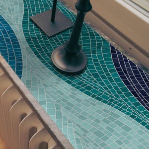Può includere: Un piano di lavoro in mosaico con un design a flusso in tonalità di verde acqua, turchese e blu navy. Il piano di lavoro è accanto a un radiatore bianco e un pavimento in legno. Una lampada nera è sul piano di lavoro.