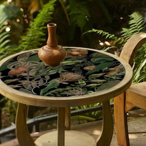Puede incluir: Una silla de madera y una mesa redonda con una tapa de mesa de estampado floral se encuentran en un patio rodeado de exuberante vegetación.