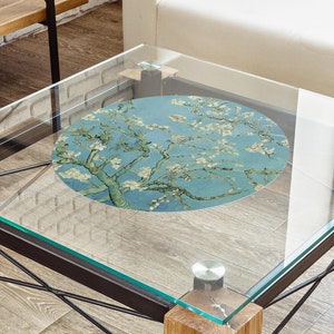 May include: A glass coffee table with a black metal frame and wooden legs. The table has a round, blue and white floral print coaster on top.