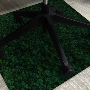 Puede incluir: Una alfombrilla verde para silla de oficina con un diseño de hojas. La alfombrilla está debajo de la base negra de una silla de oficina. Las patas y las ruedas de la silla son visibles, descansando sobre la alfombrilla. El diseño de la alfombrilla se asemeja a un suelo forestal denso.
