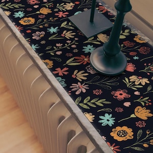May include: A black table runner with a floral pattern in shades of pink, yellow, blue, and green. The runner is decorated with a hand-drawn style and features a variety of flowers and leaves.