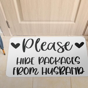 May include: White rectangular doormat with black text that reads "Please Hide Packages From Husband" with a heart on each side of the word "Please". Black boots and white sneakers are next to the mat.