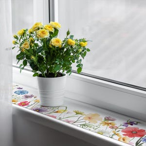 Könnte beinhalten: Ein weißes Fensterbrett mit einem Aufkleber mit Blumenmuster. Ein weißer Topf mit einem gelben Rosenstrauch steht auf dem Fensterbrett.