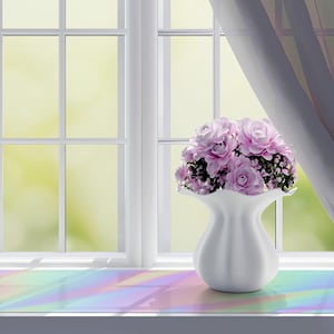 May include: A white vase filled with pink flowers sits on a windowsill in front of a window. The window has a white frame and a view of green foliage. Sheer white curtains are draped to the side. The windowsill has a rainbow-coloured surface.