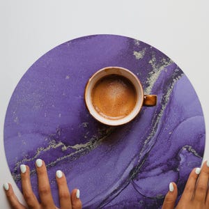May include: A top-down view of a cup of coffee on a purple and silver marbled coaster. The coffee cup is brown with a handle. The coaster is round and has a marbled design. The image also shows two hands with white nail polish.