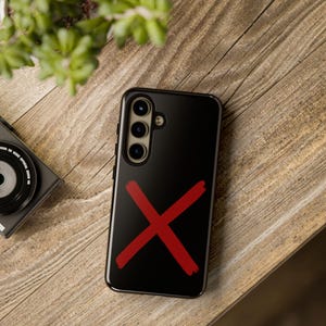 May include: Black smartphone case with a bold red "X" design. The case features a triple-lens camera and a sleek, glossy finish. The phone case is displayed on a wooden surface, with a camera and plant in the background.