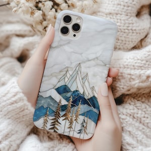 May include: A white marble phone case with a mountain design. The case features blue and gold mountain peaks and trees. The phone case is held in a person's hands.