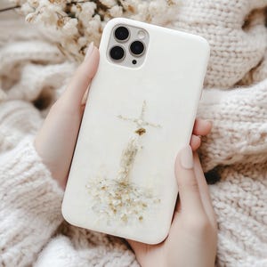 May include: A white phone case featuring a cross design made of dried flowers. The case has a glossy finish and is held against a soft, cream-colored knitted sweater. The phone case has a triple-lens camera.