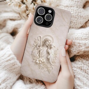 May include: A beige phone case with a marble design and a 3D relief of a religious figure holding a staff and a lamb. The case has a camera cutout and is held by a person.