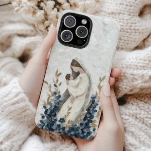 Puede incluir: Funda de teléfono con una ilustración en acuarela de una figura religiosa sosteniendo a un bebé, rodeada de flores azules y follaje. La funda tiene un fondo blanco texturizado y un módulo de cámara negro.