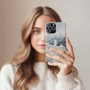 May include: A phone case featuring a painting of a person in a white robe standing among sheep, overlooking a mountainous landscape. The case has a light gray and blue color scheme, with a glossy finish. The phone case is held in front of a person.