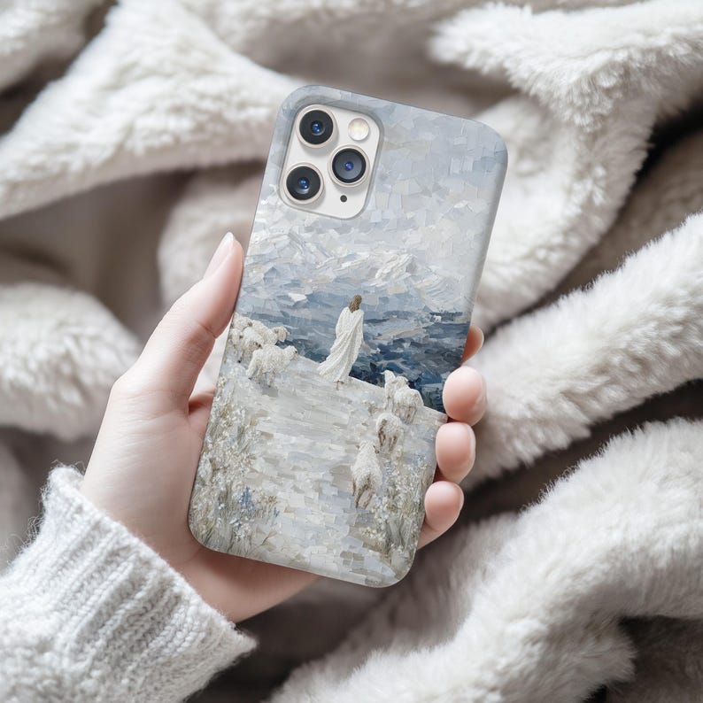 May include: A phone case featuring a painting of a figure in white robes with sheep, set against a backdrop of mountains and a cloudy sky. The artwork is rendered in cool tones of blue, gray, and white.