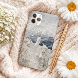 May include: A phone case featuring a painting of a figure in white robes with sheep, overlooking a mountainous landscape. The case is light blue and white, with a gold pen and white flowers nearby.