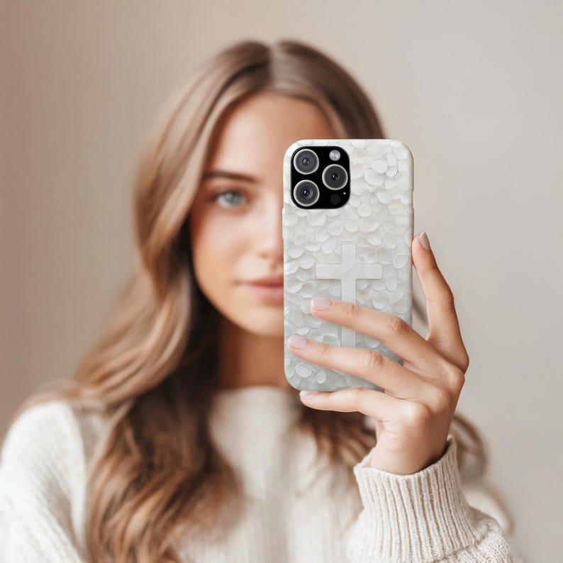 May include: White phone case with a textured pattern and a raised cross design. The case has a triple-lens camera cutout. The phone case is held in front of a person.