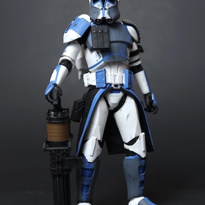 May include: A posable action figure of a soldier wearing white and blue armor with a weathered finish. The figure wears a helmet with a blue visor, a black chest pouch, and a black fabric kama. It holds a large black and brown rotary blaster cannon.