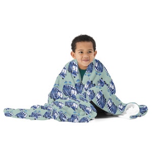 Puede incluir: Una manta verde claro con un patrón repetido de furgonetas camper azules y blancas. La manta está envuelta alrededor de una persona. Las furgonetas camper tienen detalles a cuadros y a rayas. La manta es de material suave.