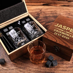 May include: A wooden gift set featuring two engraved whiskey glasses, ice stones, and tongs. The glasses are etched with "ANDREW groomsman". The box is engraved with "JASPER GROOMSMAN 11-17-2025". A glass of amber liquid and ice stones are also present.