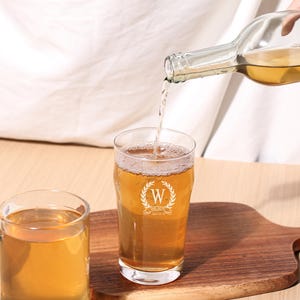 May include: A clear glass beer mug with a white monogram design featuring the letter "W" and the words "Wedding" and "Est. 2023" is being filled with a light amber liquid from a clear glass bottle.