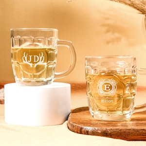May include: Two clear glass beer mugs with handles. The mug on the left has the initials "JD" and antlers etched into the glass. The mug on the right has the letter "E" in a decorative frame and the name "Elliot" etched into the glass.