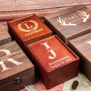 May include: Several wooden boxes, each with a unique engraved design and name. The boxes are various shades of brown, with different fonts and decorative elements, including a laurel wreath and antlers. Some boxes have dates and titles.