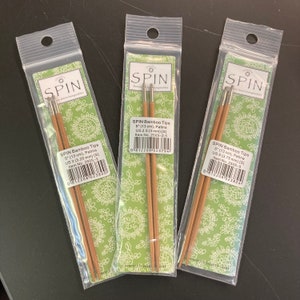 May include: Three packs of bamboo knitting needles with silver tips. The packs are labelled "SPIN Bamboo Tips" and list the size of the needles in US and mm. The needles are 5 inches (13 cm) long.