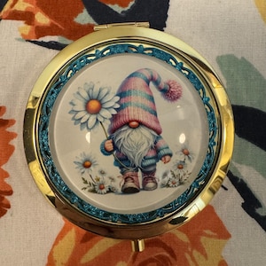 May include: A round, gold-toned compact mirror with a teal border. The mirror's face features a whimsical illustration of a garden gnome holding a daisy. The gnome wears a striped pink and blue hat and sweater.