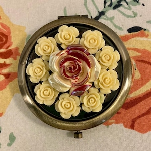 May include: A round, antique-style compact mirror with a bronze-colored frame. The mirror's face is decorated with cream-colored rosebuds surrounding a larger rose with red accents. The compact is closed.