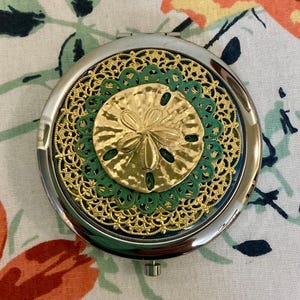May include: A round, silver-toned compact mirror with a gold-colored sand dollar design. The sand dollar is surrounded by a green and gold filigree pattern. The mirror is closed and has a small button to open it.