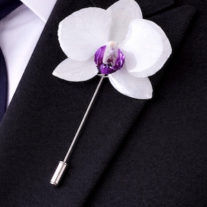 May include: A white orchid flower lapel pin with a purple center, attached to a silver pin. The pin is displayed on a black suit jacket, with a white collared shirt and a navy blue tie visible in the background.