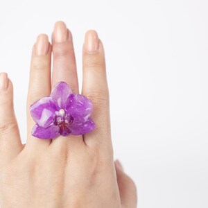 May include: A purple orchid flower ring. The ring is on a person's finger.