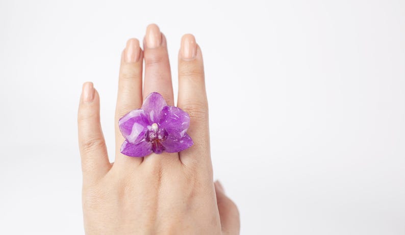 May include: A purple orchid flower ring. The ring is on a person's finger.