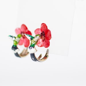 Könnte beinhalten: Ein Paar Creolen mit einem Kolibri-Design. Die Ohrringe haben eine goldfarbene Oberfläche mit einem schwarzen, weißen, grünen und blauen Kolibri und einer roten Blume. Auf weißem Hintergrund.