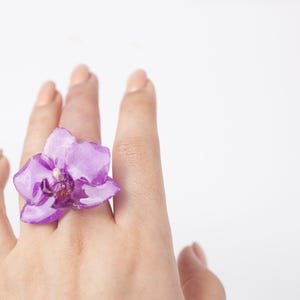 May include: A hand wearing a ring with a purple orchid flower.