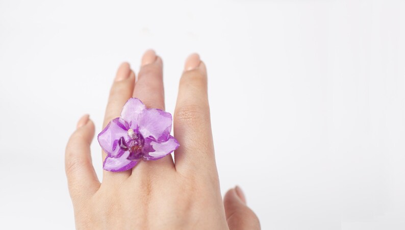 May include: A hand wearing a ring with a purple orchid flower.