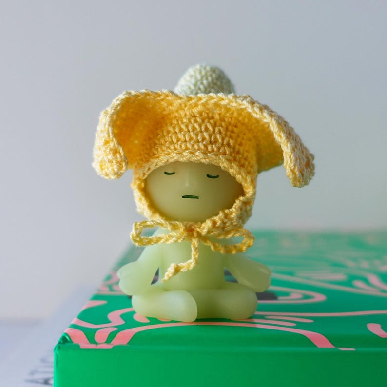 May include: A small, green, glow-in-the-dark figurine wearing a yellow crocheted hat with floppy ears. The figurine is sitting in a meditative pose on a green and pink surface.