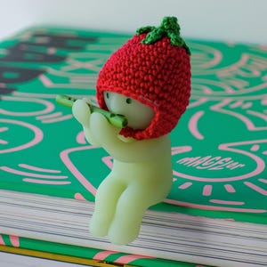 May include: A small, green, glow-in-the-dark figurine wearing a red crocheted strawberry hat plays a green flute. The figurine is sitting on a green book with pink and white markings. The book has the word "MAGIC" printed on it.